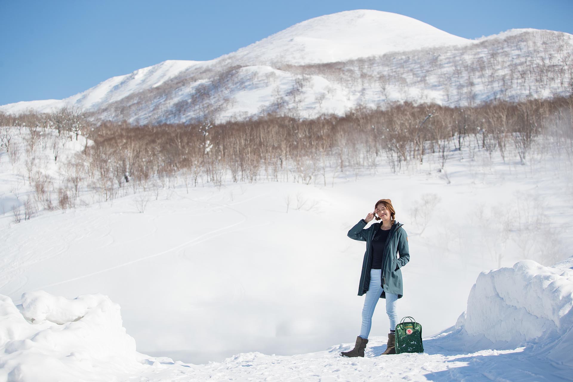 Woman with a backpack posing in deep snow with Mount Niseko Annupuri rising under a clear blue sky behind her