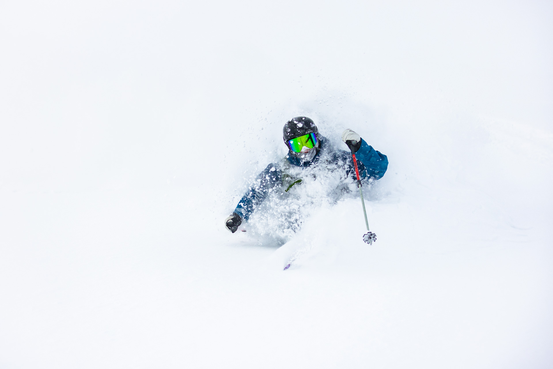Skier in a blue jacket and green goggles carving through deep powder snow in Niseko, Japan, engulfed in a white snow spray