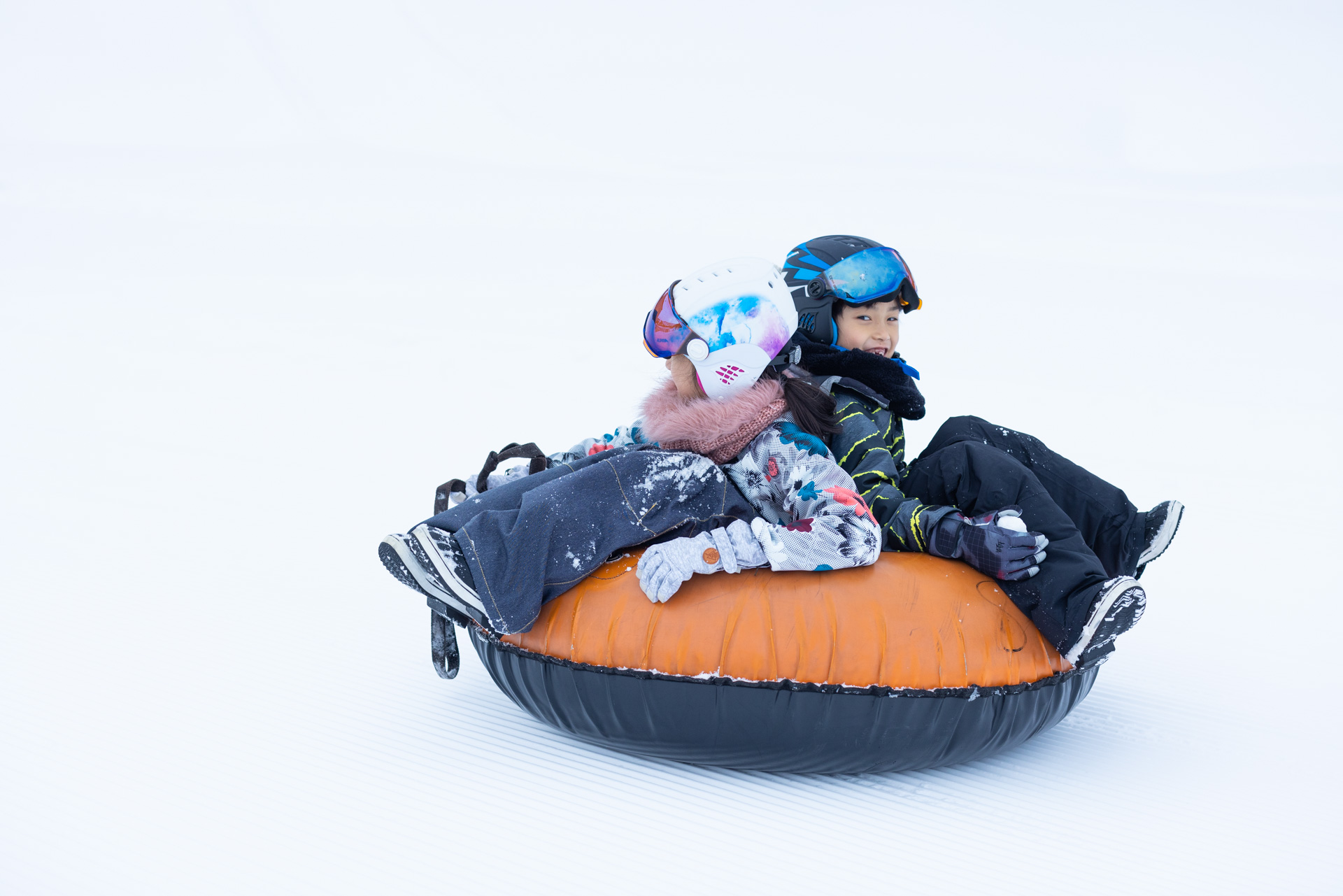 Two children in helmets and ski gear laughing while riding an orange snow tube down a snowy slope in Niseko