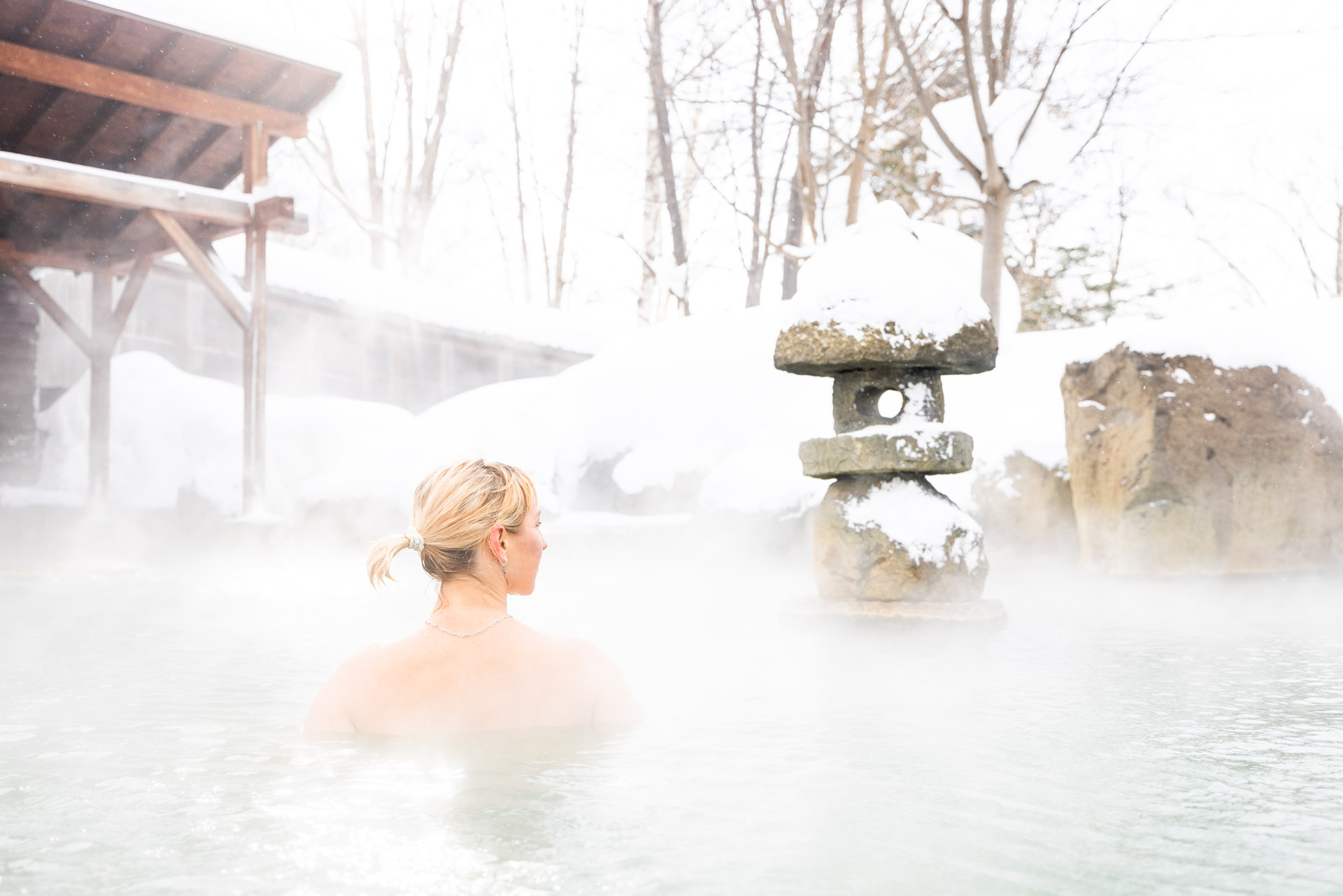 Woman soaking in a steaming outdoor onsen in Niseko with a snow-dusted stone lantern and winter trees in the mist
