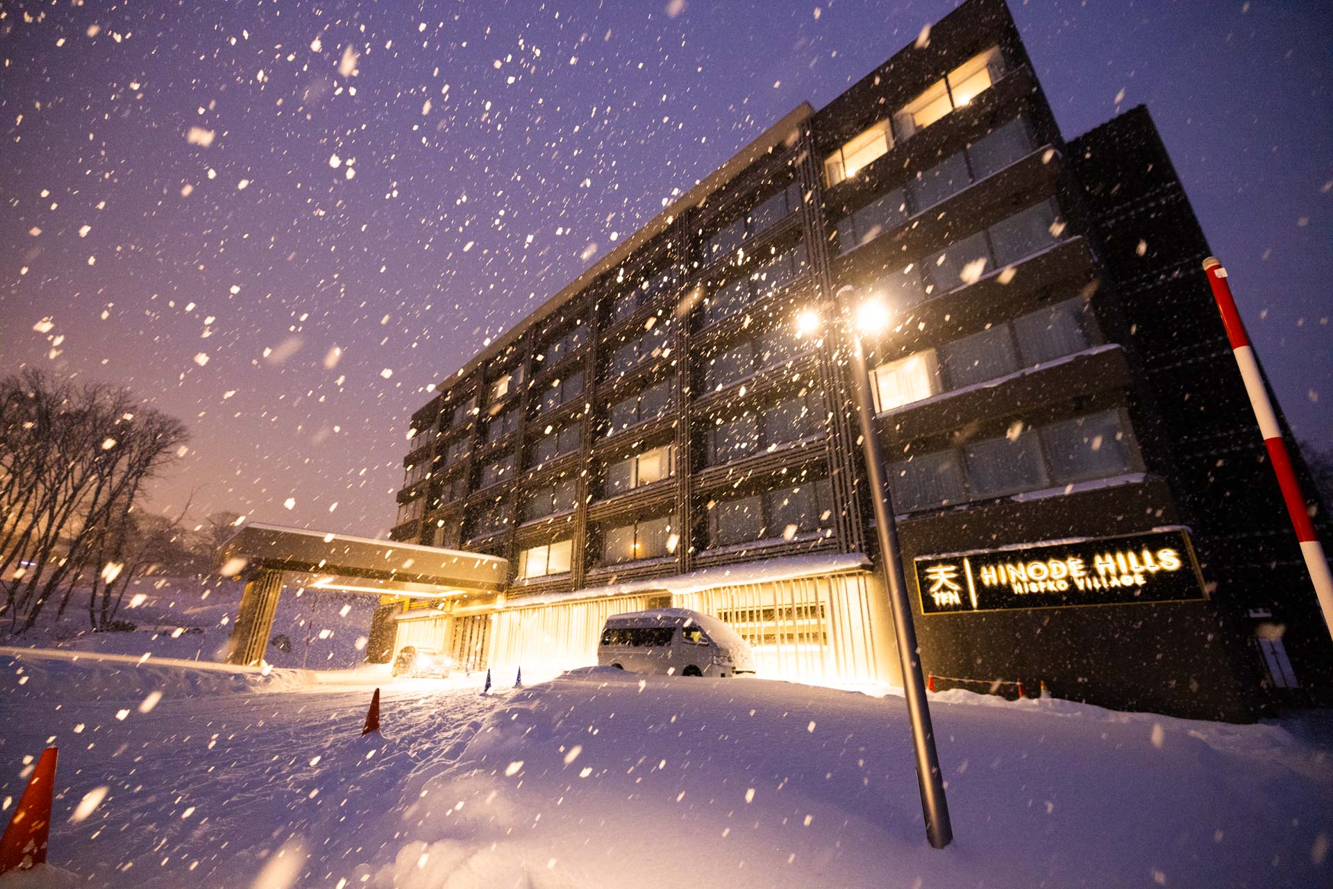 Hinode Hills Niseko Village hotel exterior at night during heavy snowfall, with warm glowing lights and deep powder snow drifts