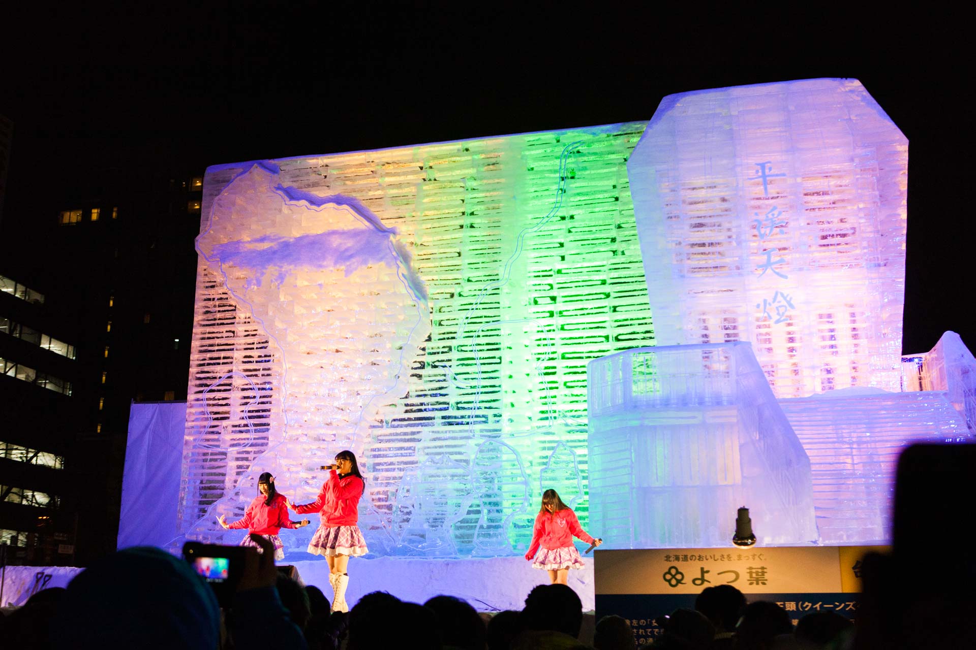 J-pop performers on stage in front of a massive illuminated ice sculpture at the Sapporo Snow Festival at night