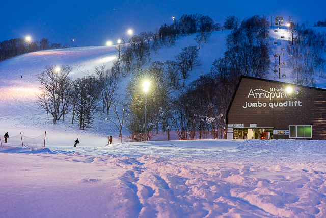Night skiing at Niseko Annupuri with illuminated slopes, the Jumbo Quad Lift station, and skiers descending under bright lights.
