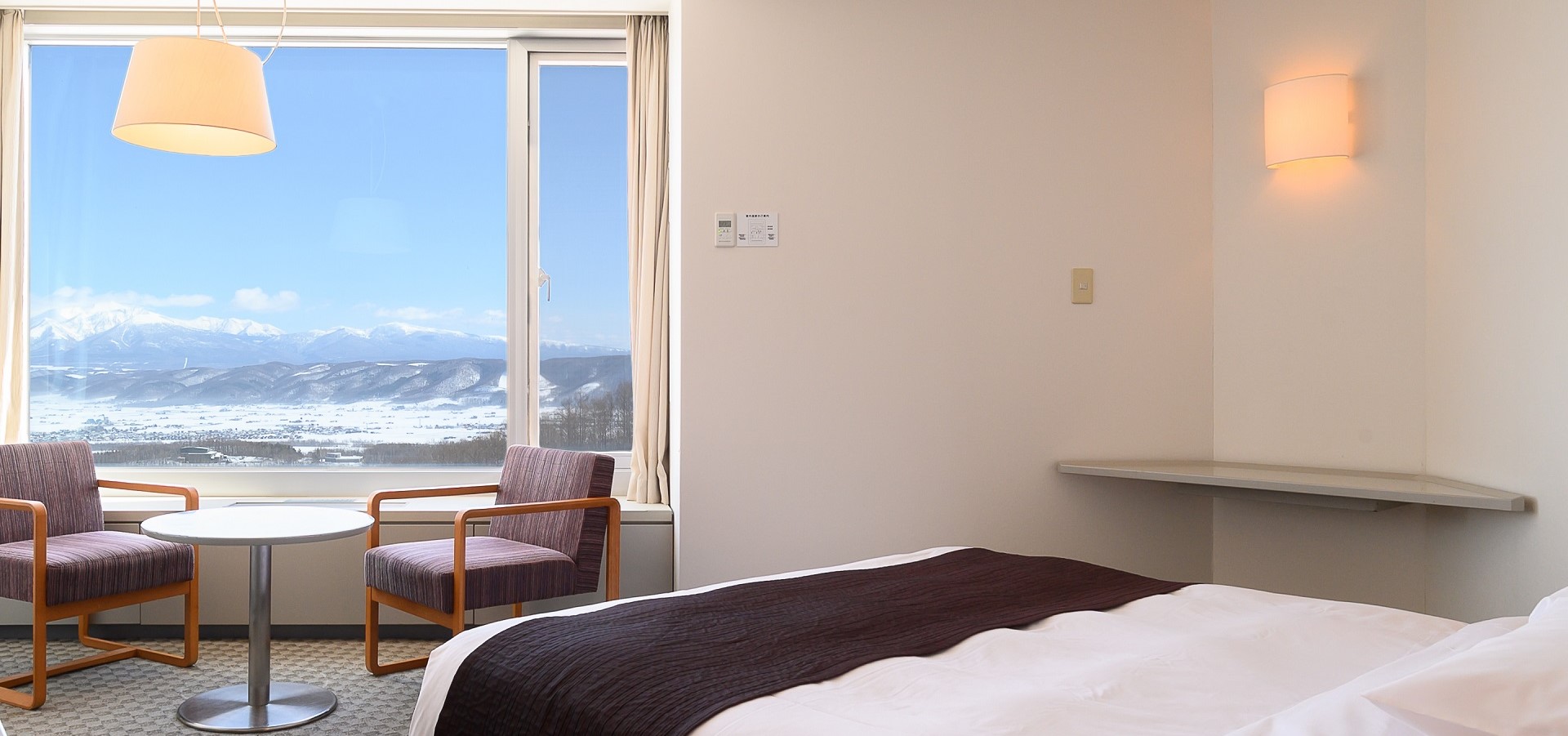 Hotel room with mountain and snowy valley views, large window, cozy seating area, and modern bed in Furano, Japan