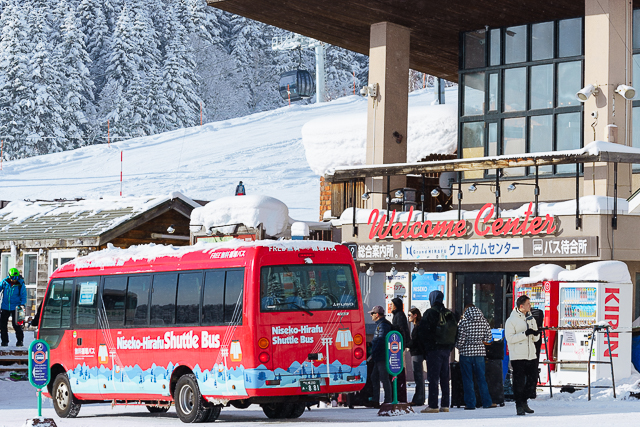 Red Niseko-Hirafu Shuttle Bus parked at the Welcome Center with skiers and snow-covered slopes in the background.