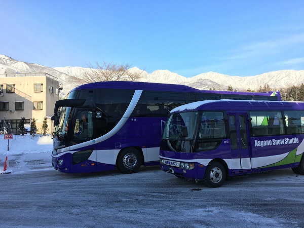 Two purple Nagano Snow Shuttle buses parked in a snowy lot with mountains in the background.