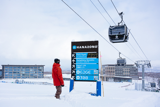 Skier in a red jacket looking at a resort signpost with the Hanazono Symphony Gondola and Park Hyatt Niseko in the background.