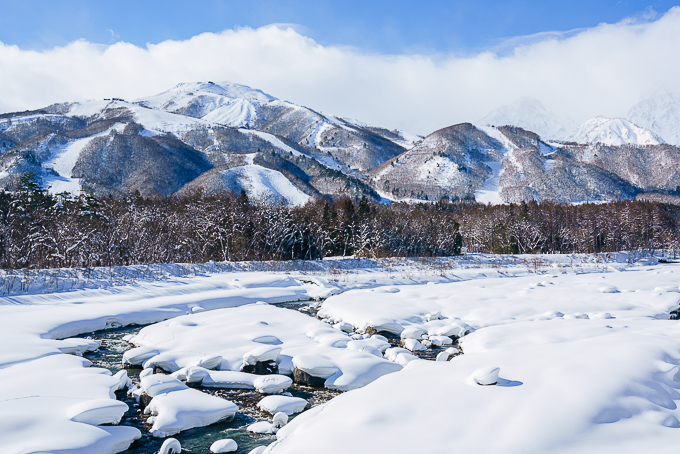 Snowy riverbank and winter forest with the snow-capped Hakuba mountains and Happo-One ski slopes under a blue sky.