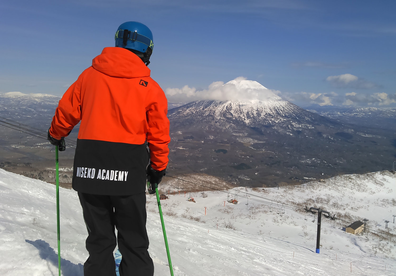 A skier in an orange Niseko Academy jacket looks out over a vast valley and snow-capped Mount Yotei.