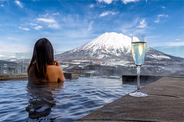 Woman in an infinity onsen bath with a glass of champagne overlooking a snowy Mount Yotei landscape.