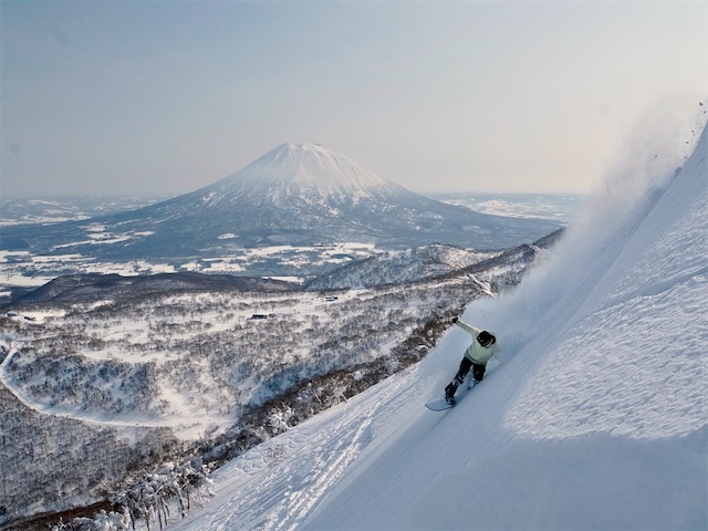 Snowboarder riding through deep powder on a steep slope with a panoramic view of Mount Yotei.