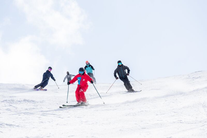A ski instructor in a bright red suit leads a group of students down a bright, sunlit groomed run.
