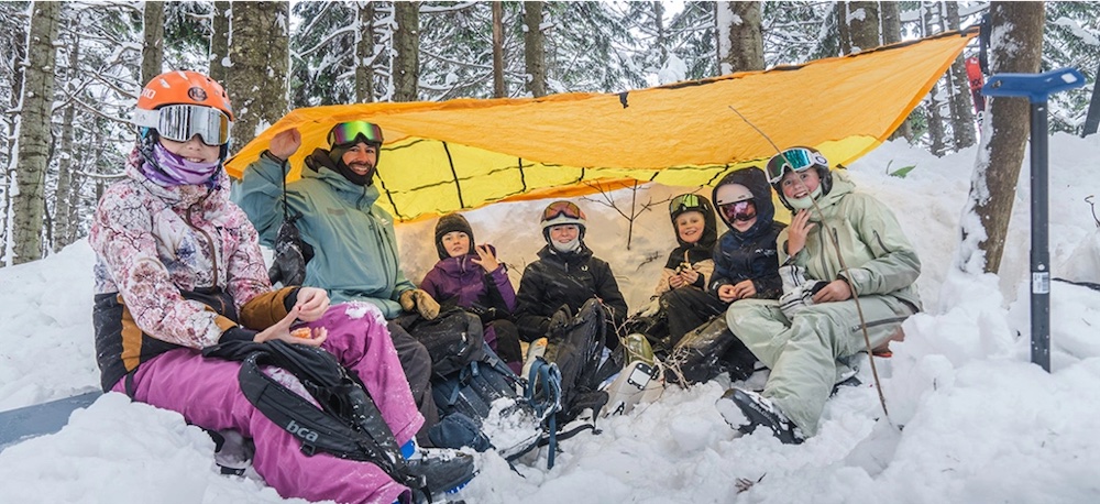 A group of skiers huddling under a yellow tarp within a forest.