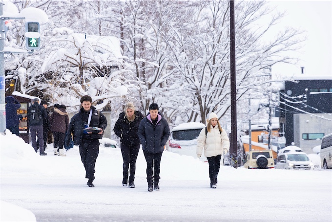 Tourists in winter jackets walk across a snow-covered street in a Japanese ski village during winter.