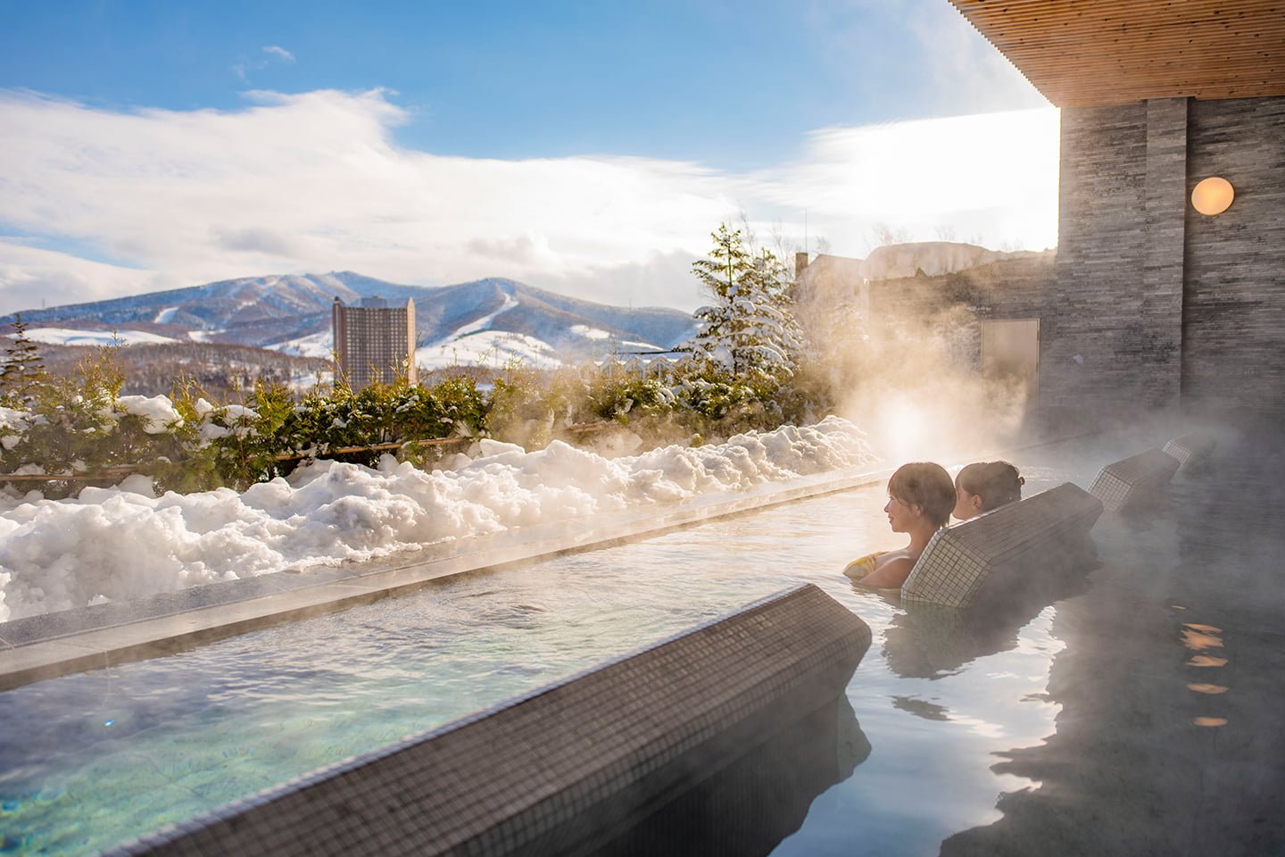 People relaxing in a steaming outdoor hot spring onsen overlooking snow-covered mountains in Rusutsu.