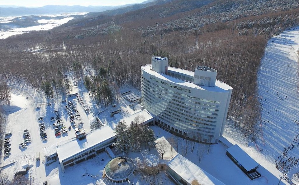 Aerial view of the curved New Furano Prince Hotel surrounded by snow, pine forests, and ski runs.