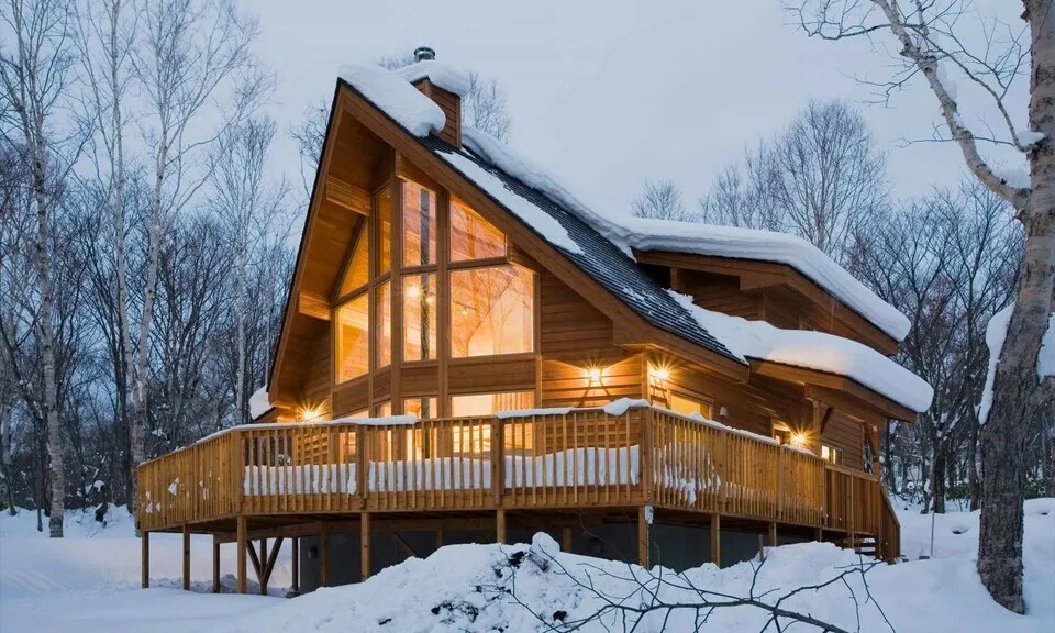 A-frame wooden cabin glowing at night, surrounded by snow-covered trees and a large deck.
