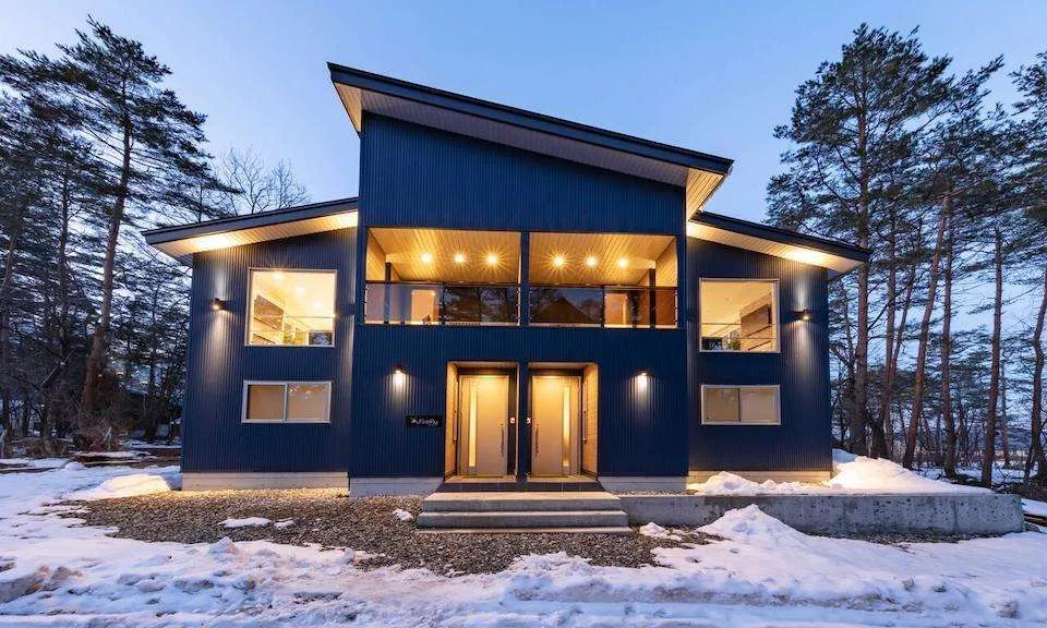 Modern dark blue duplex with illuminated balconies and glowing windows in a snowy forest at dusk.