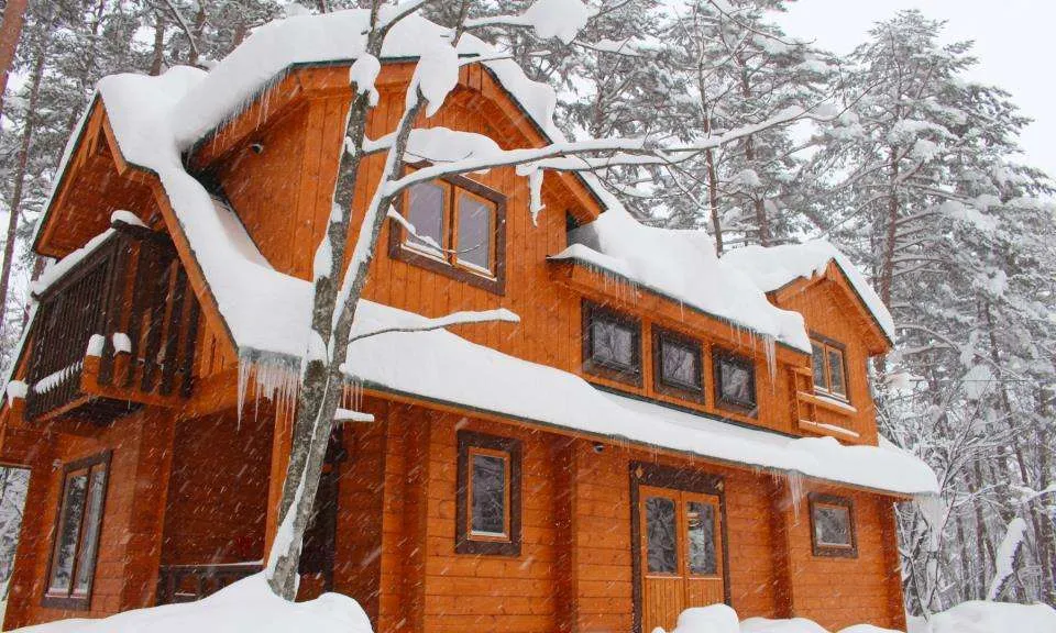 Traditional two-story orange log cabin covered in deep snow and icicles among pine trees.