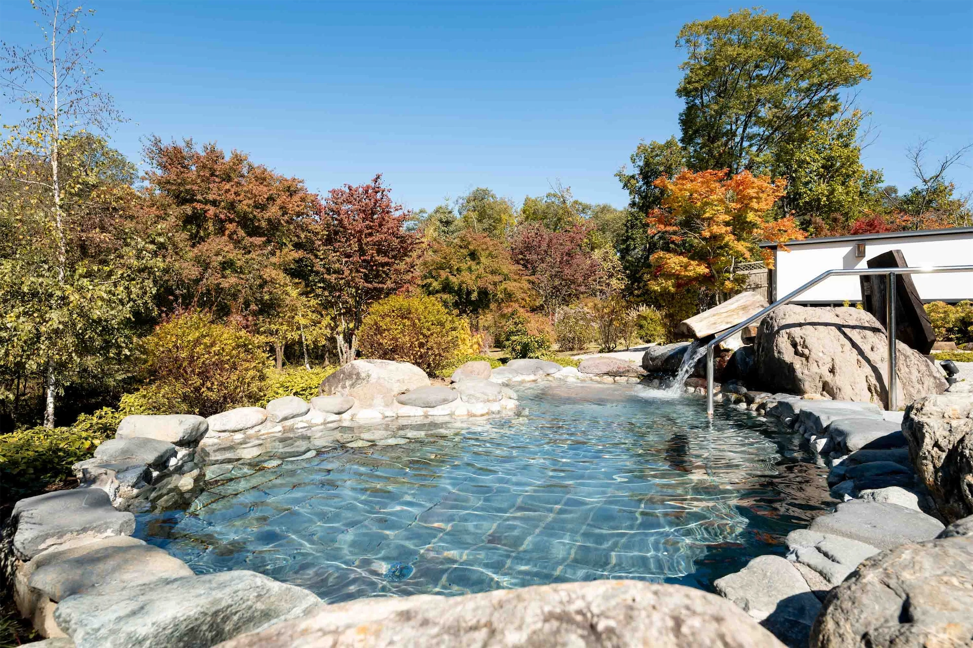 Outdoor rock onsen bath in Hakuba surrounded by colorful autumn trees and a clear blue sky.