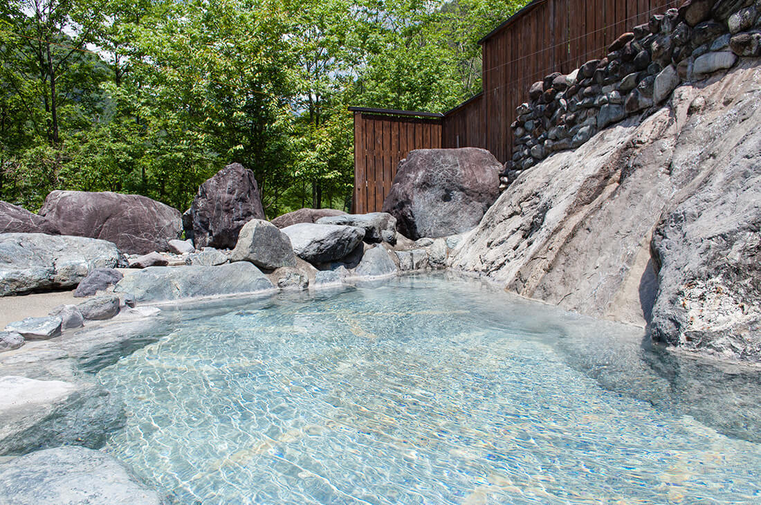 Natural outdoor rock onsen bath with clear water surrounded by lush green trees in summer.