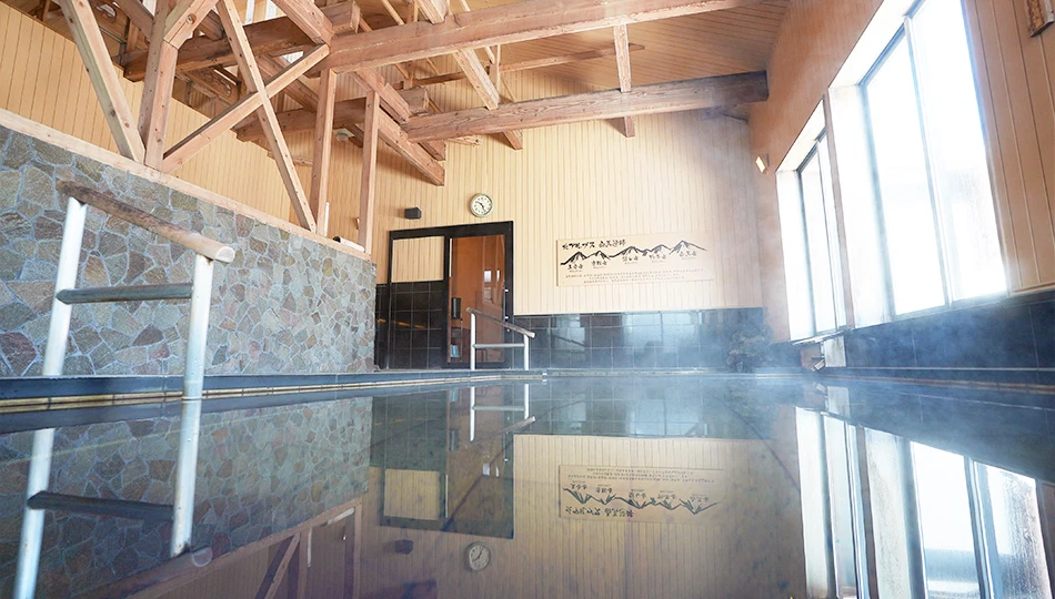 Indoor onsen bath with stone walls, exposed wooden ceiling beams, and large bright windows.