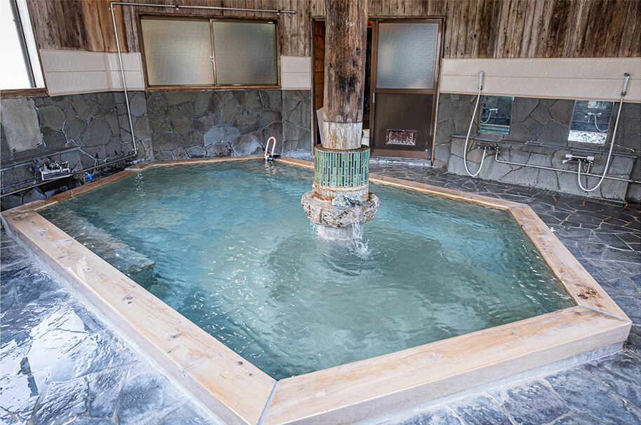 Indoor hexagonal onsen bath with stone walls, wooden trim, and a central pillar fountain.