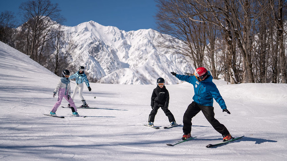 A ski instructor in a blue jacket teaches a family on a sunny slope with jagged alpine peaks behind.