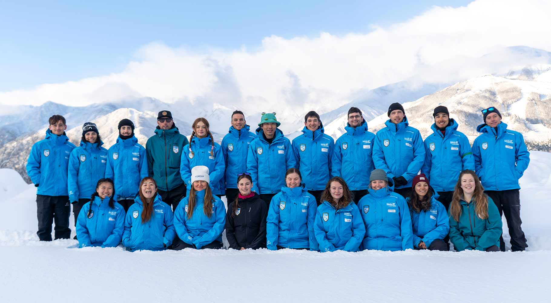 A large group of ski instructors in matching blue jackets pose together in the snow against a mountain backdrop.