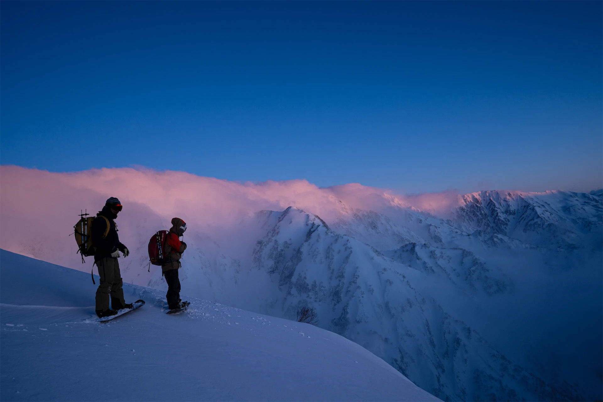Two backcountry skiers stand on a snowy ridge overlooking pink-tinted clouds and mountain peaks in Hakuba at sunset.