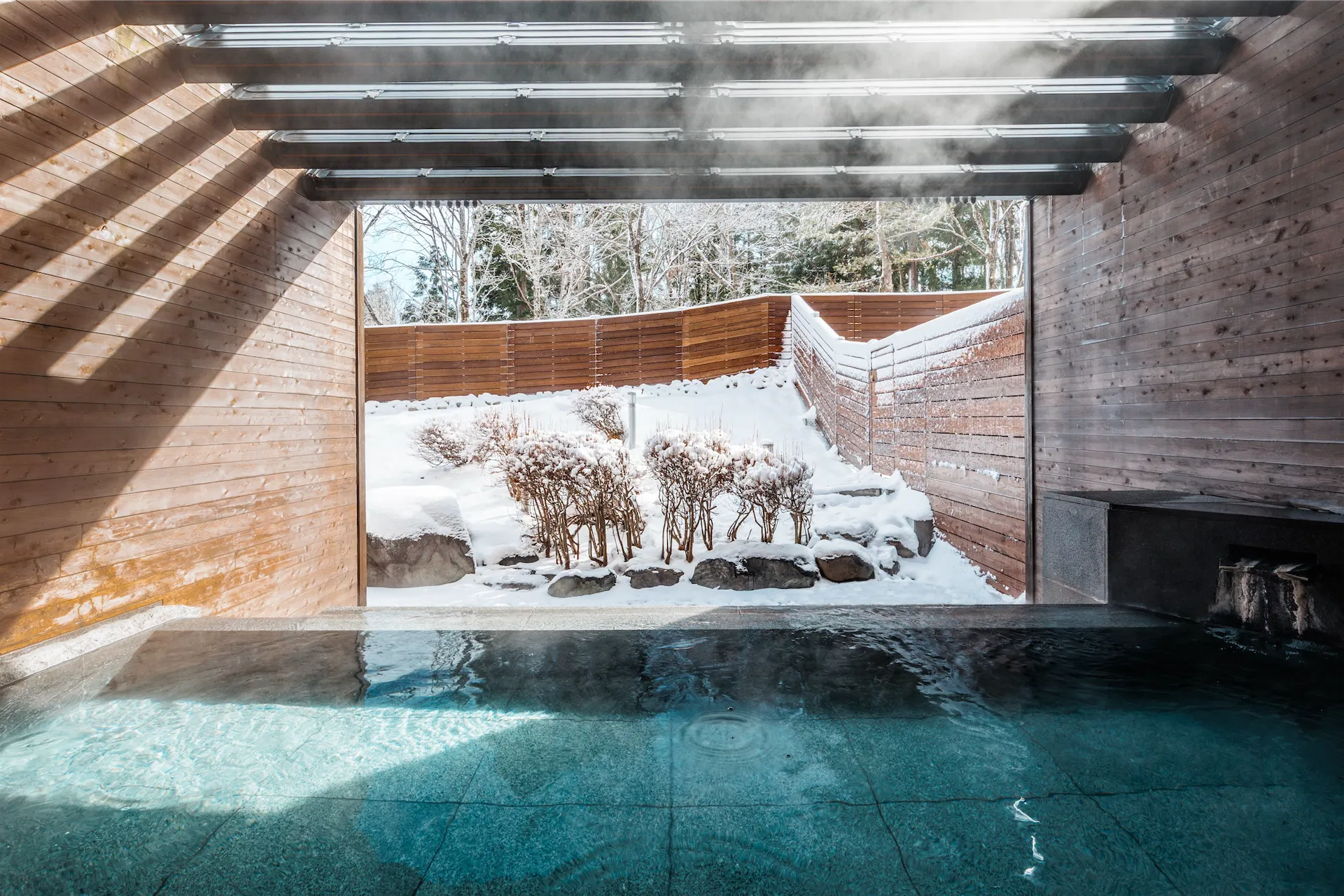 Modern semi-outdoor onsen at Hakuba Tokyu Hotel with steam rising over a snowy garden view.