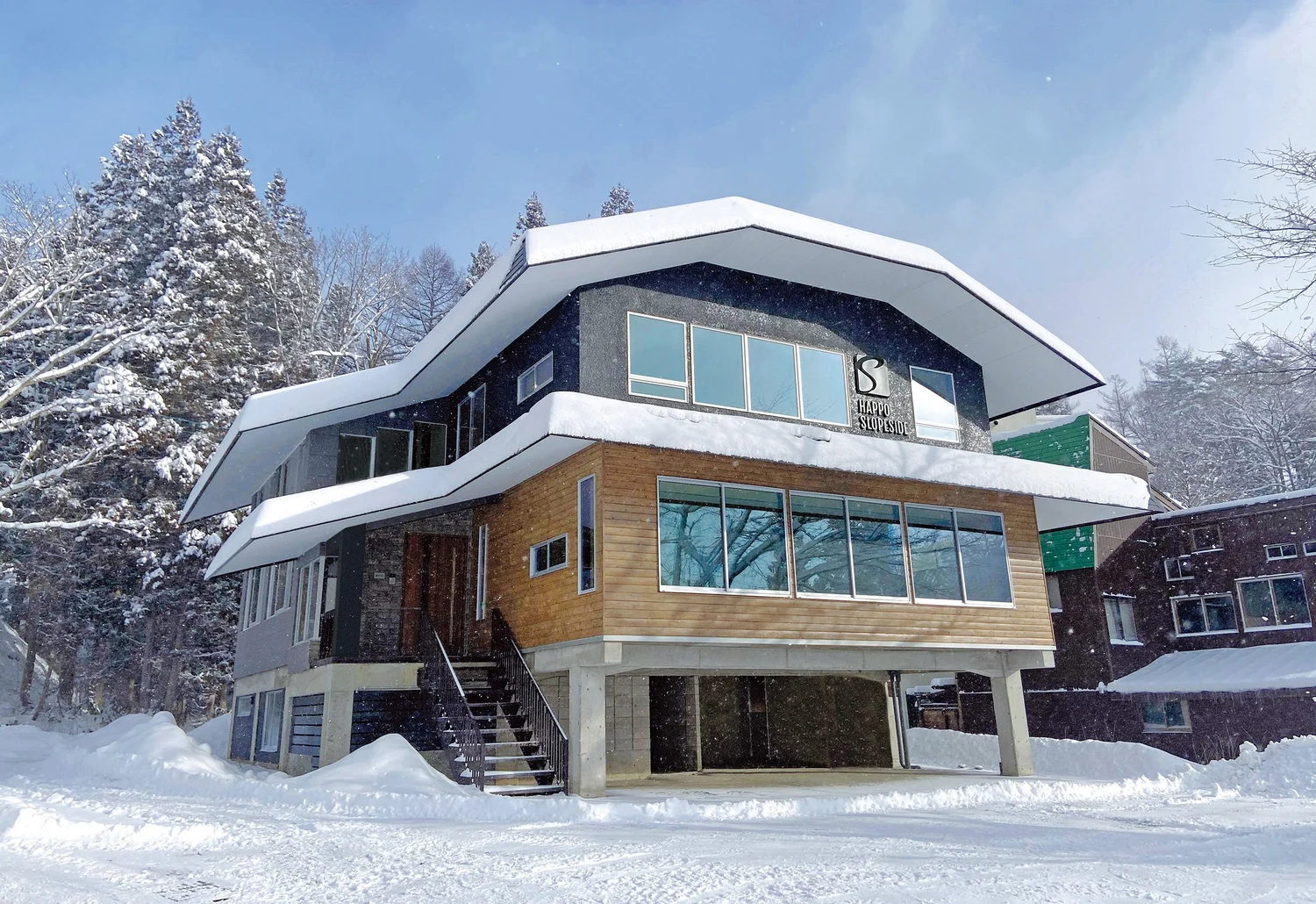 Snow-covered mountain lodge with wood and charcoal siding featuring large windows and an elevated entrance.