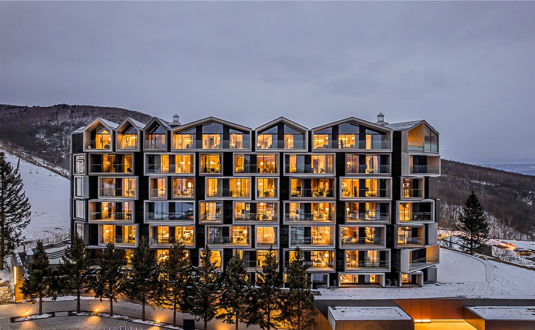 Contemporary glass-fronted apartment building illuminated at dusk against a snowy hillside.