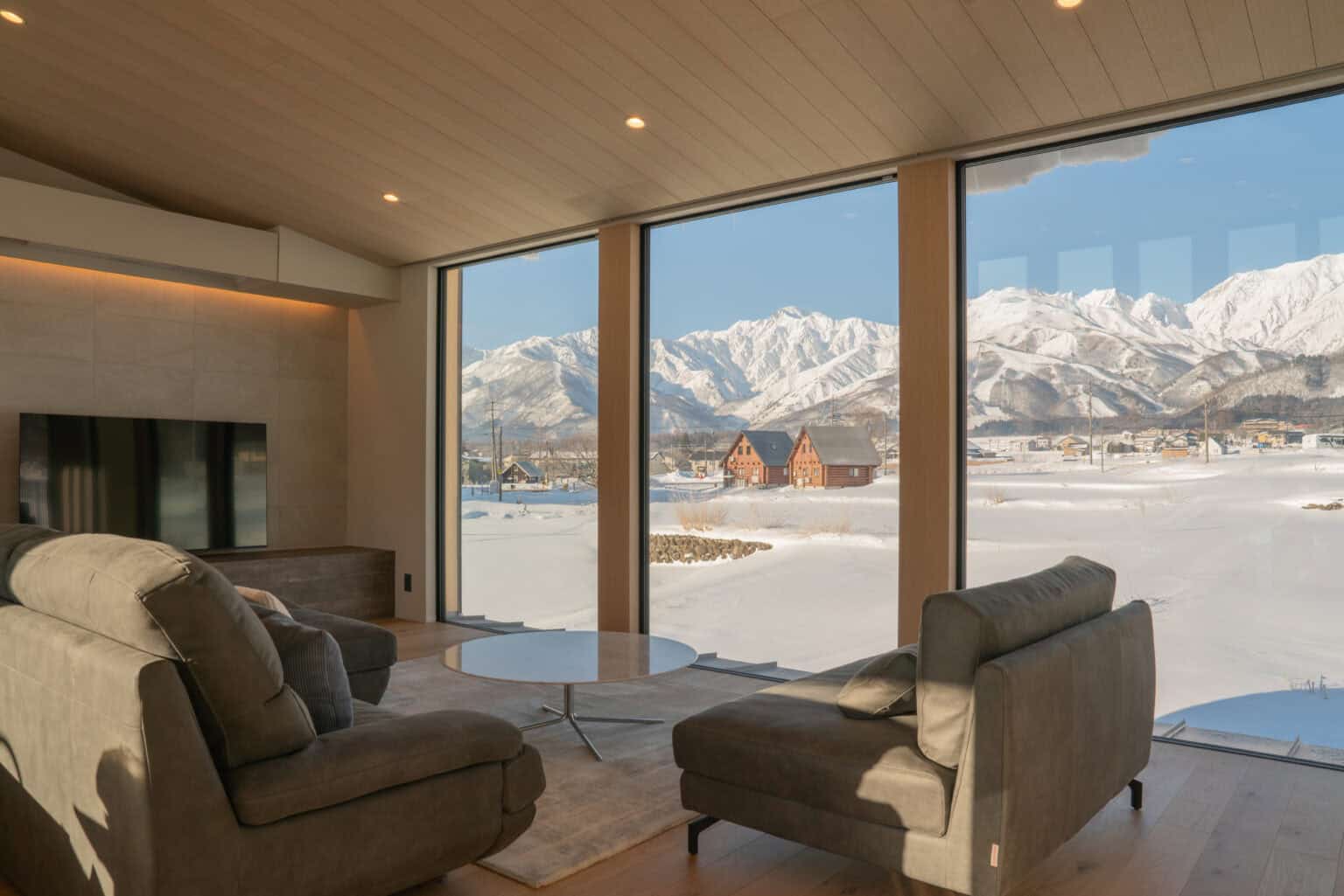 Minimalist living room with grey sofas and large windows framing a view of snowy fields and mountains.