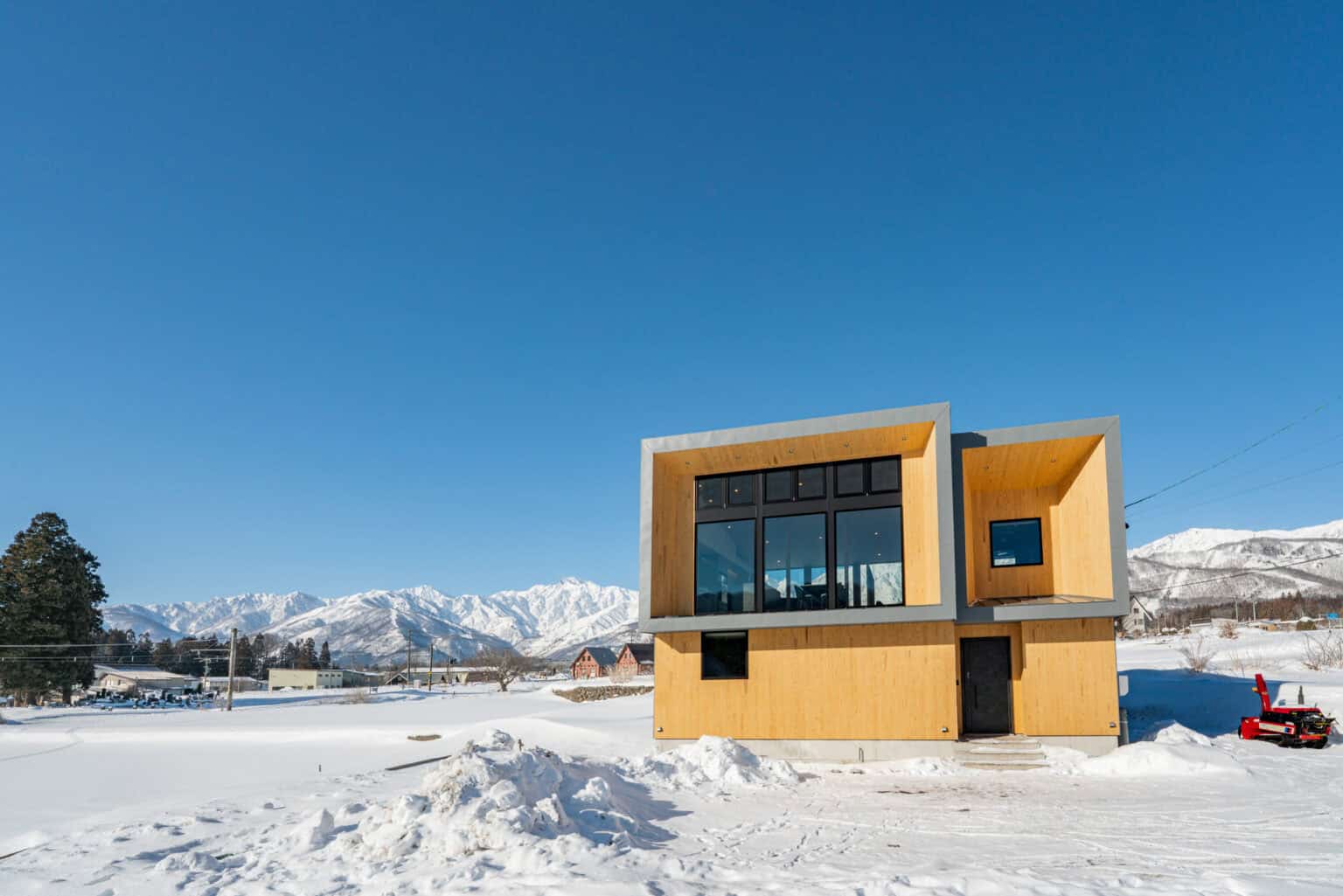 Modern box-style timber house with large windows in a snowy landscape against snow-capped mountains.