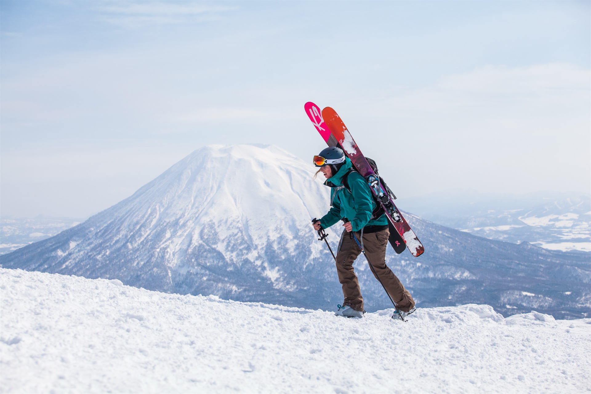 Skier hiking a snowy ridge in Niseko with skis on her backpack and a view of Mount Yotei in the background.