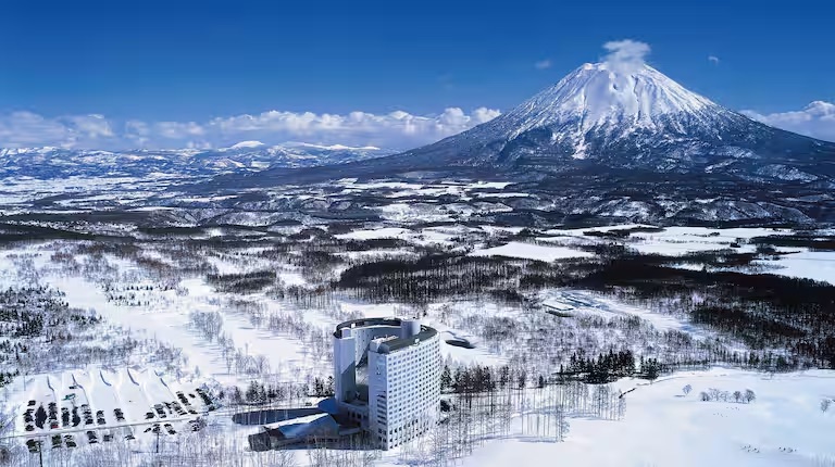Aerial view of a curved high-rise hotel in a snowy landscape with Mount Yotei in the distance.