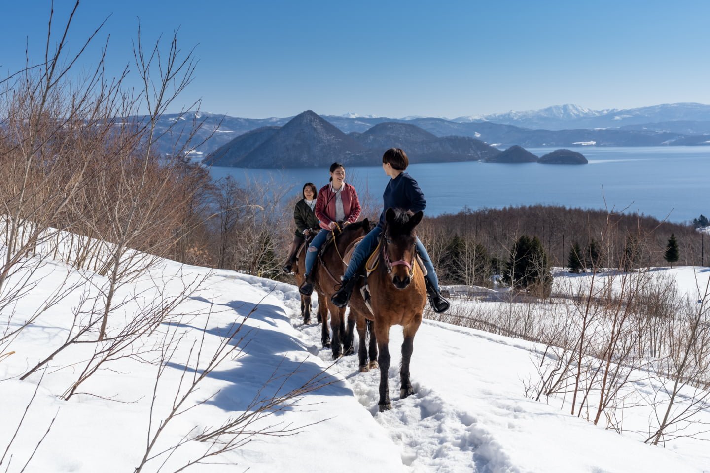 Three people enjoying a guided horseback ride through a snowy trail, overlooking the stunning blue waters of Lake Toya and its central islands under a clear sky.