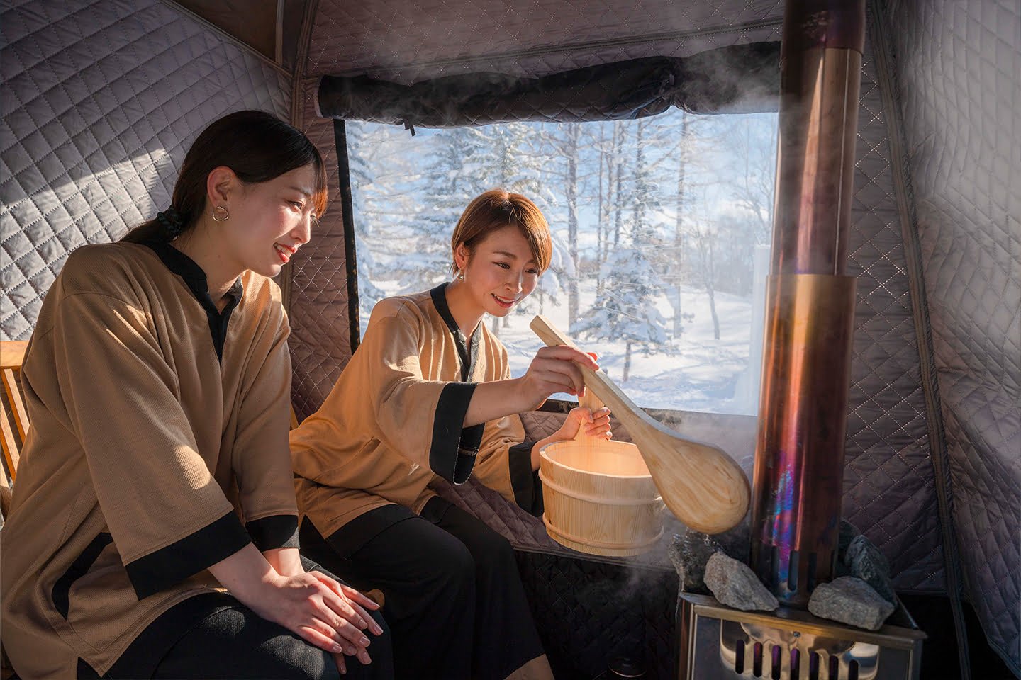 Two Japanese women enjoy a steaming sauna in a cozy tent, looking out at a snowy forest view.