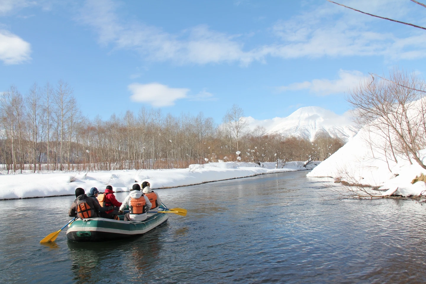 A group in a blue raft navigating a calm, snowy river landscape during winter in Rusutsu.