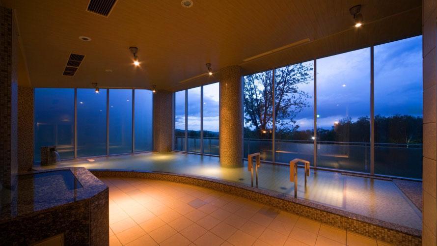 A modern indoor onsen with large floor-to-ceiling windows overlooking a dark forest at twilight.