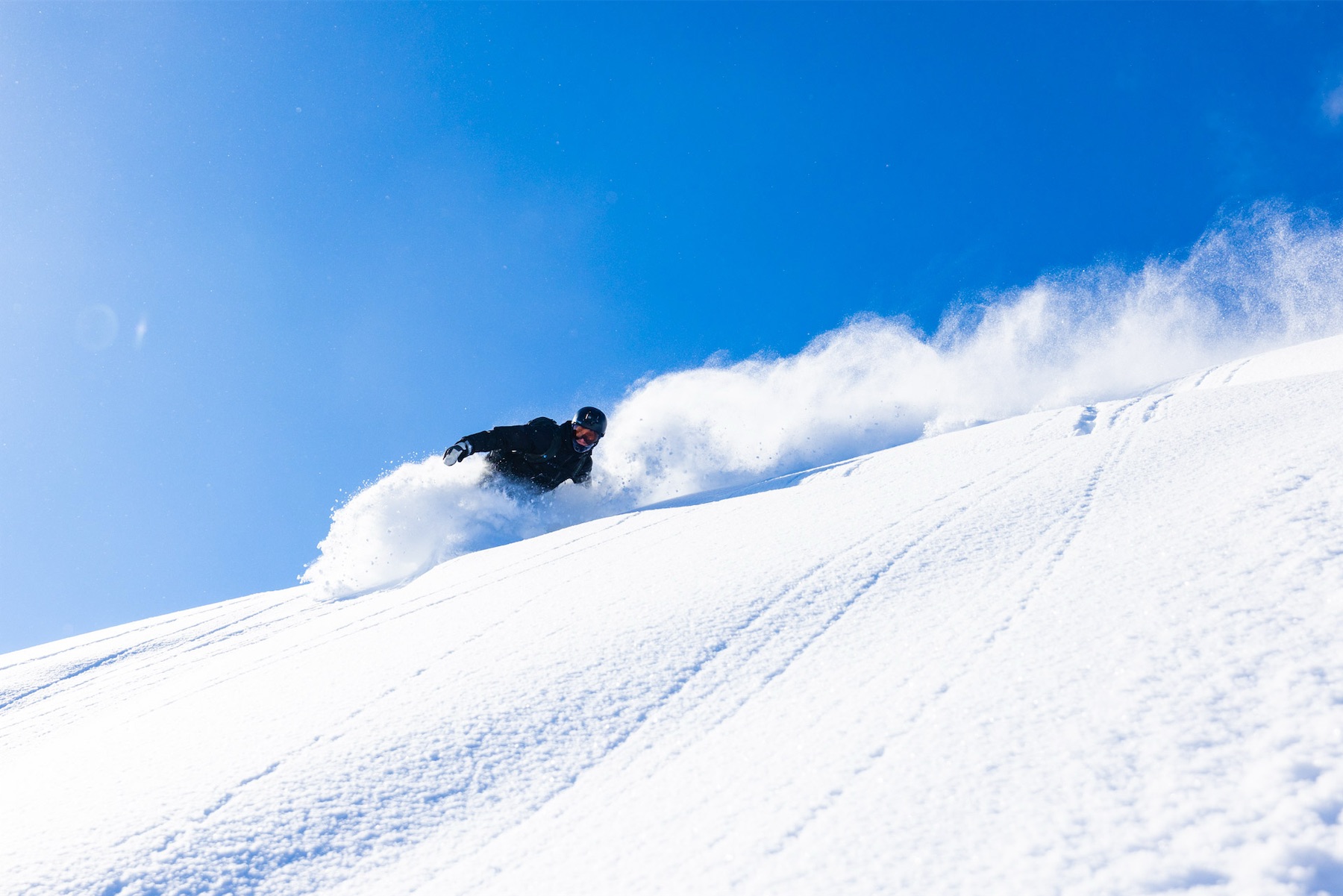 Snowboarder in black carving through deep white powder under a vibrant blue sky.