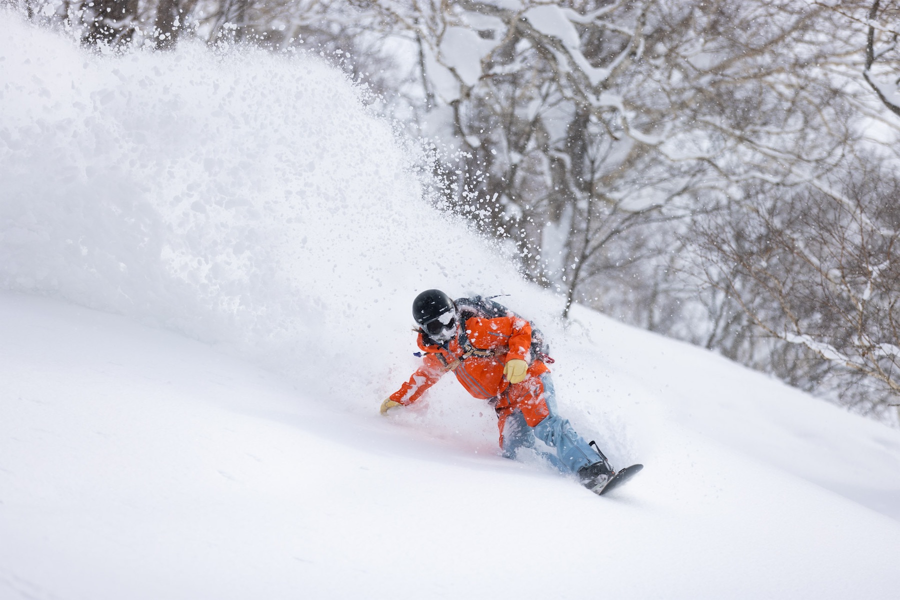 Snowboarder in an orange jacket kicking up a large spray of powder on a steep slope.