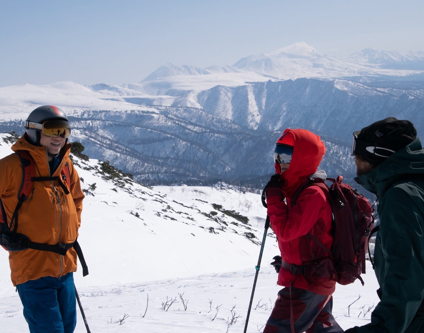 Three skiers in colorful winter gear standing on a snowy ridge overlooking mountain ranges.