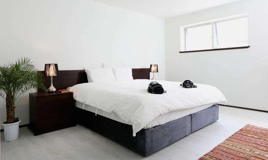 Minimalist bedroom with a large white bed, dark wood headboard, and bedside lamps against white walls.