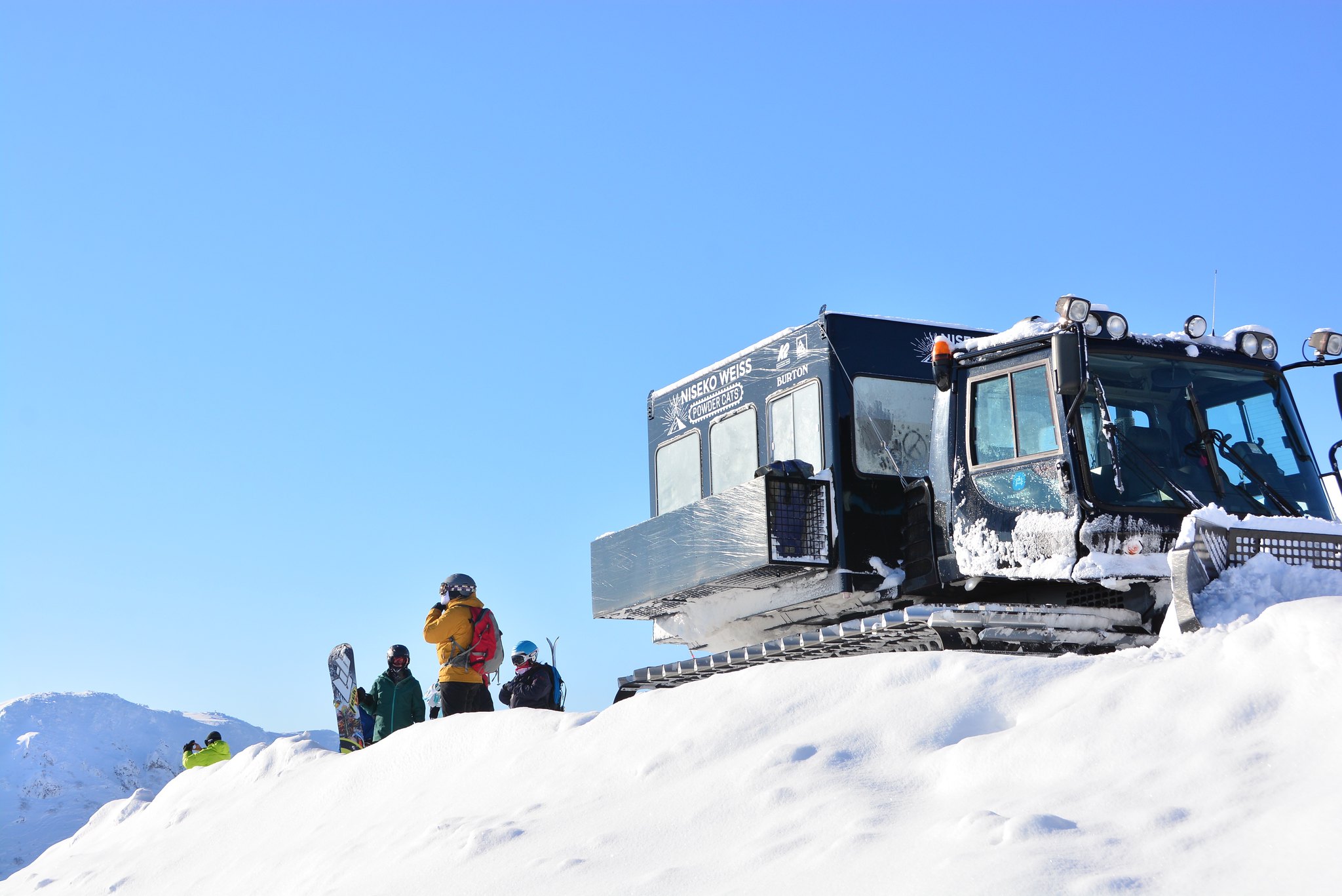 Snowboarders standing by a Niseko Weiss Powder Cats snowcat on a sunny, snow-covered mountain peak.