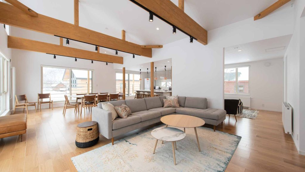 Spacious open-plan living area featuring exposed wooden beams, a gray sectional sofa, and light wood flooring.