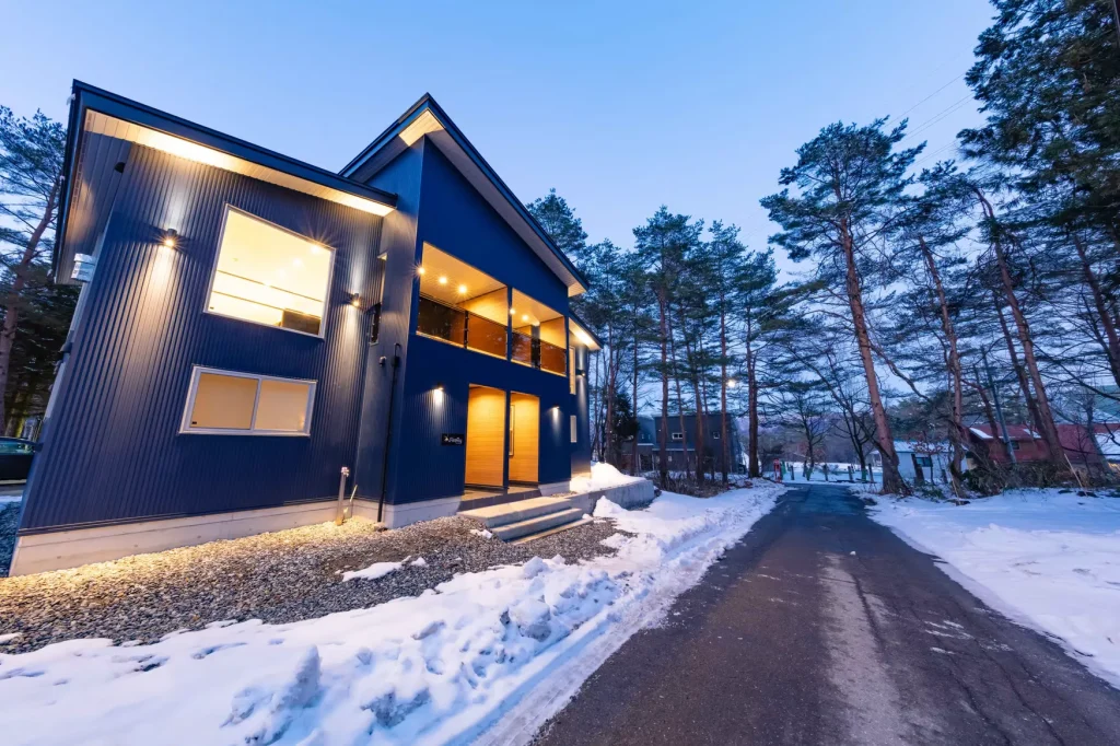 Modern dark blue chalet with warm interior lights on a snowy road at dusk.