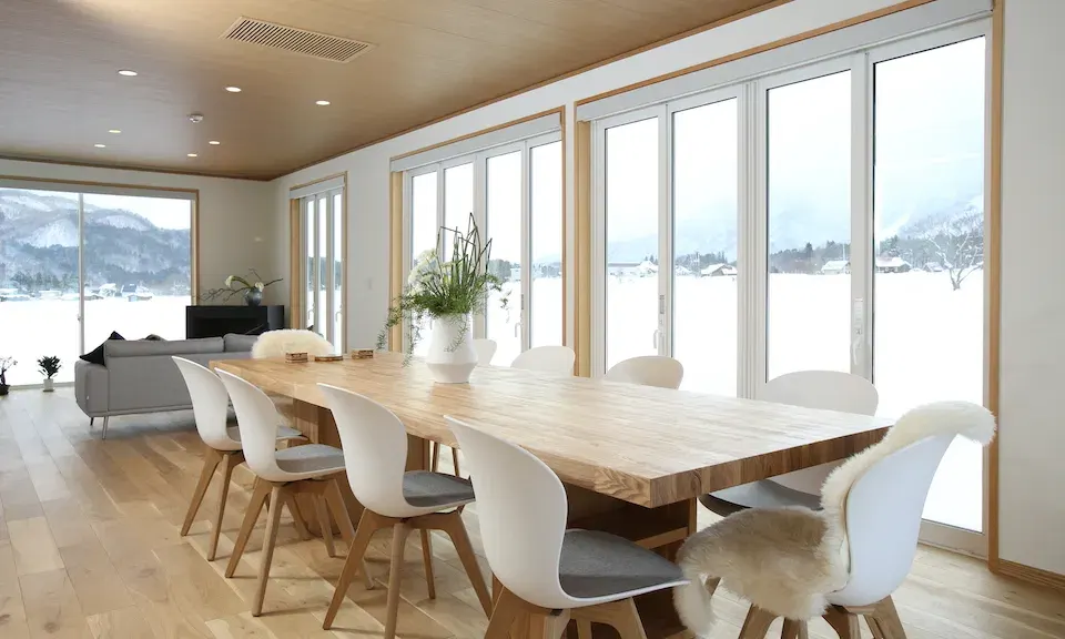 Bright dining space with a long wooden table, white chairs, and expansive windows facing snowy fields.