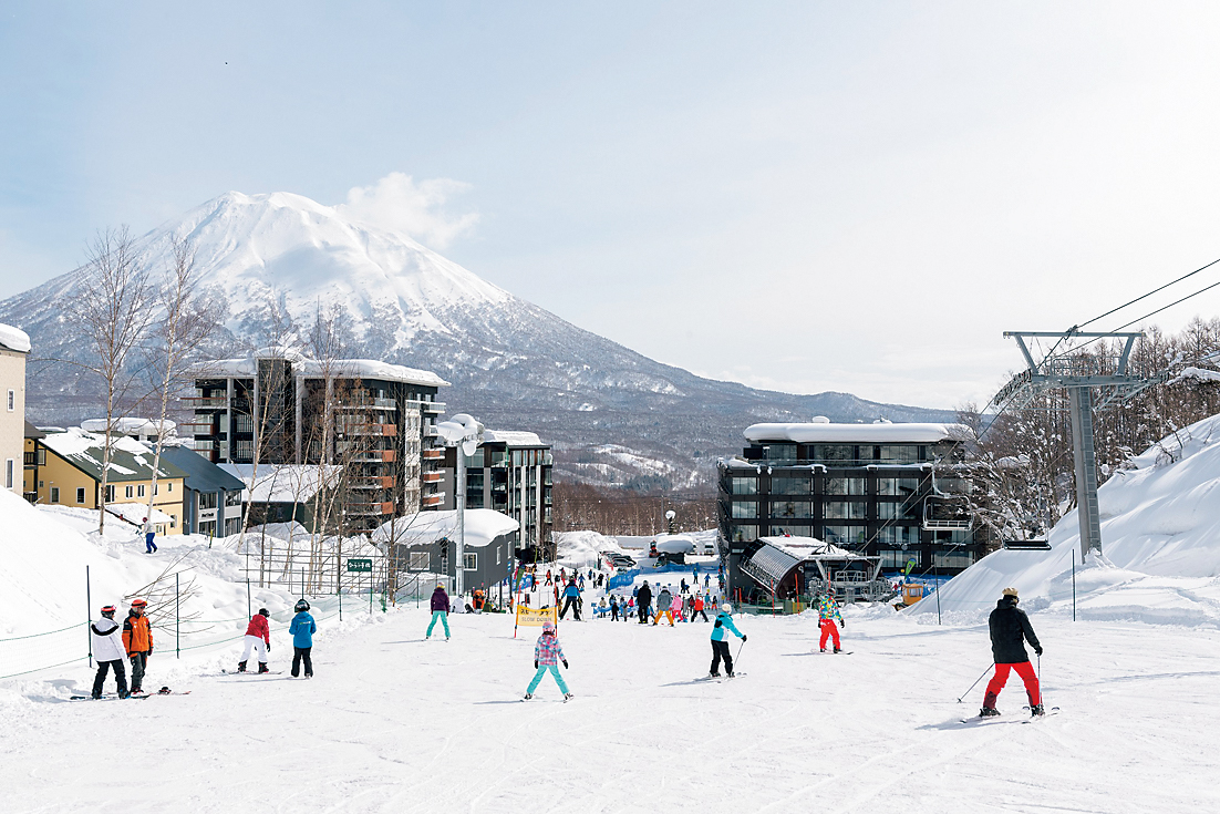 People skiing down the slopes of Niseko ski resort towards hotels with Mount Yotei in the background.